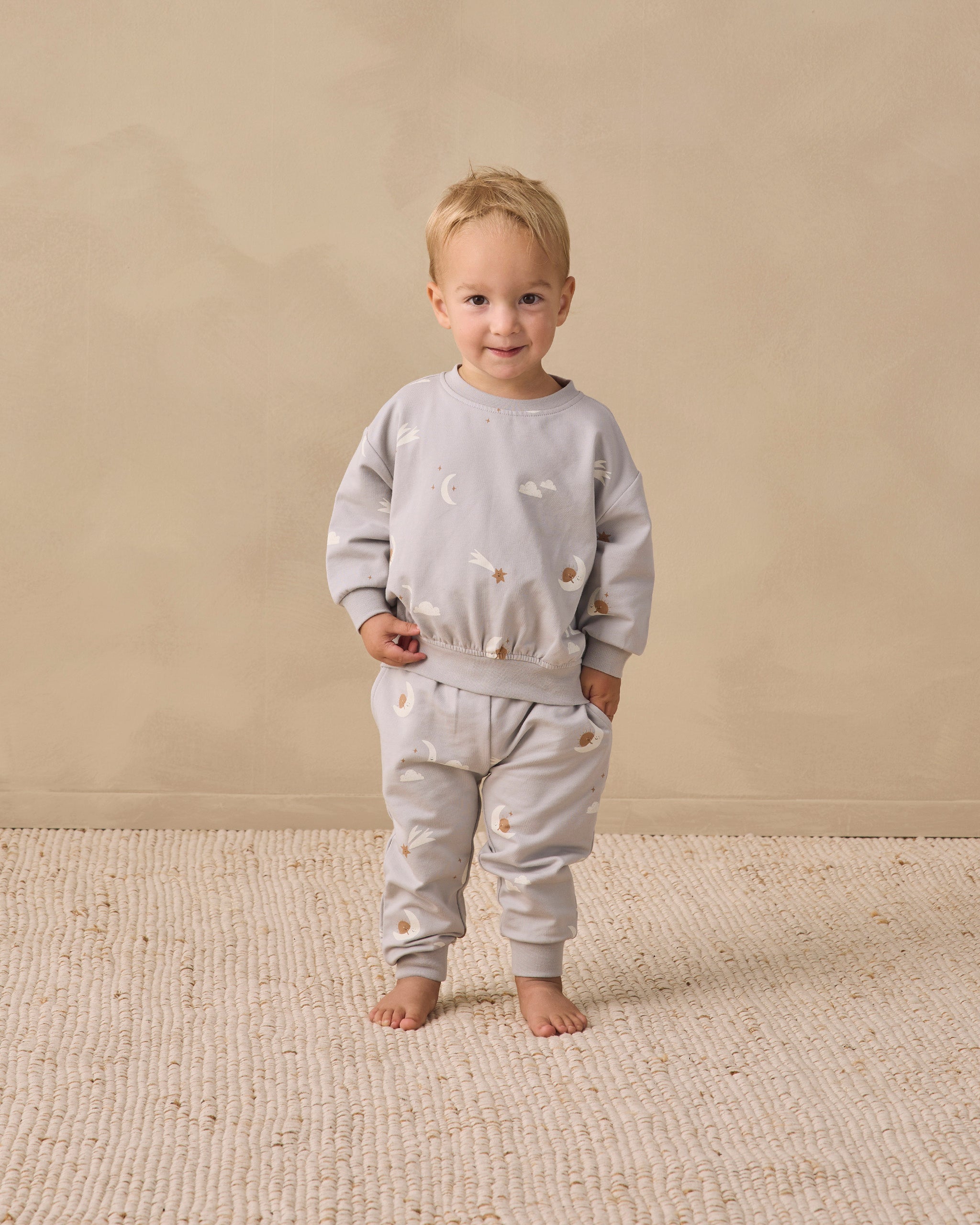 A toddler stands barefoot on a textured cream rug against a warm beige studio wall, wearing a light gray sweatshirt and matching jogger pants with small embroidered motifs.