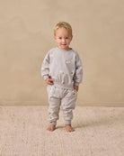 A toddler stands barefoot on a textured cream rug against a warm beige studio wall, wearing a light gray sweatshirt and matching jogger pants with small embroidered motifs.