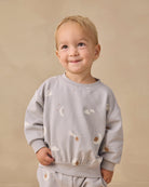 A smiling toddler with blonde hair wearing a light gray long-sleeve sweatshirt and matching pants with a celestial print of white clouds, moons, and stars, set against a plain beige background.