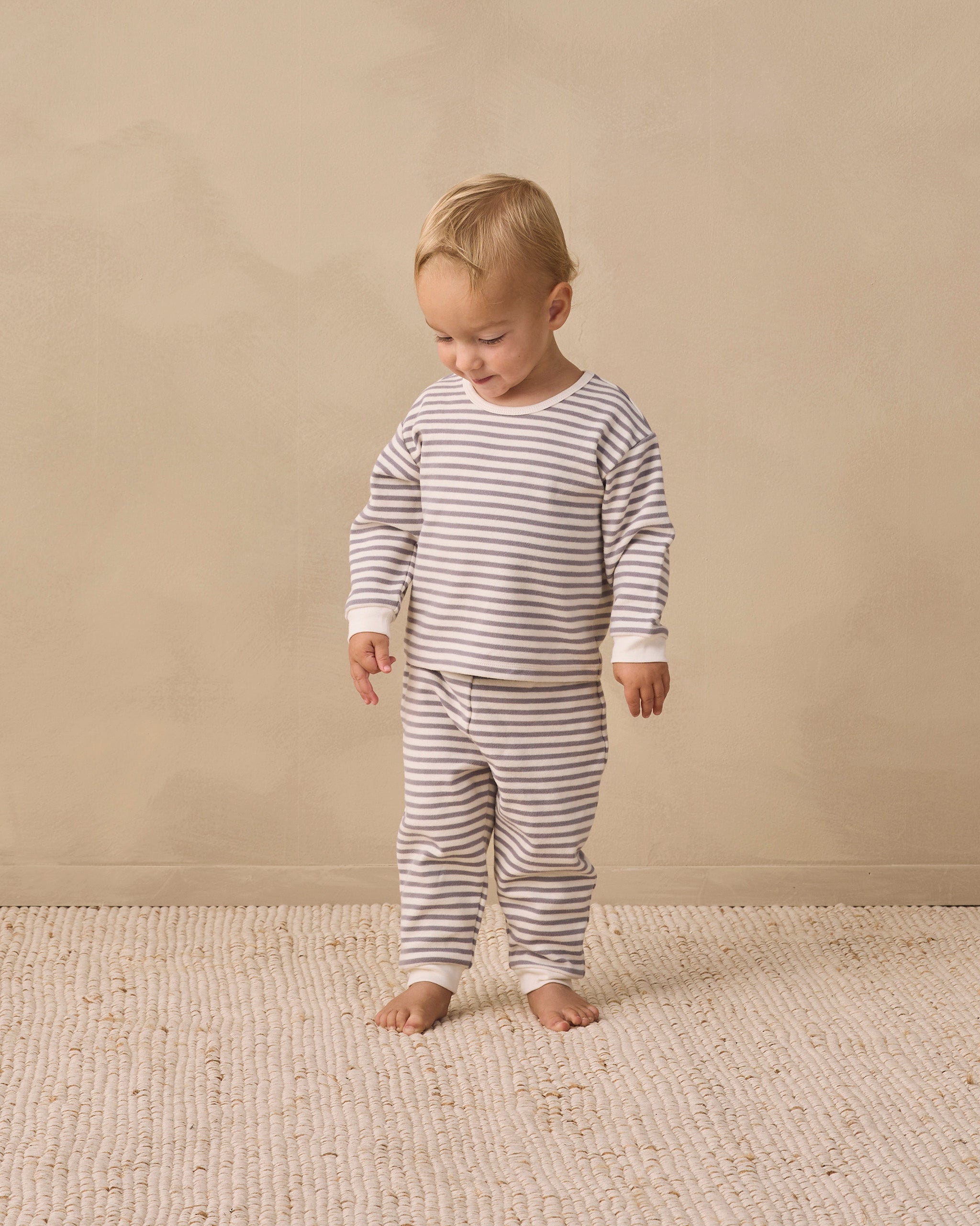 A toddler standing barefoot on a textured cream rug against a warm beige wall, wearing a long-sleeve and matching striped pajama set in beige and white.