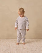 A toddler standing barefoot on a textured cream rug against a warm beige wall, wearing a long-sleeve and matching striped pajama set in beige and white.