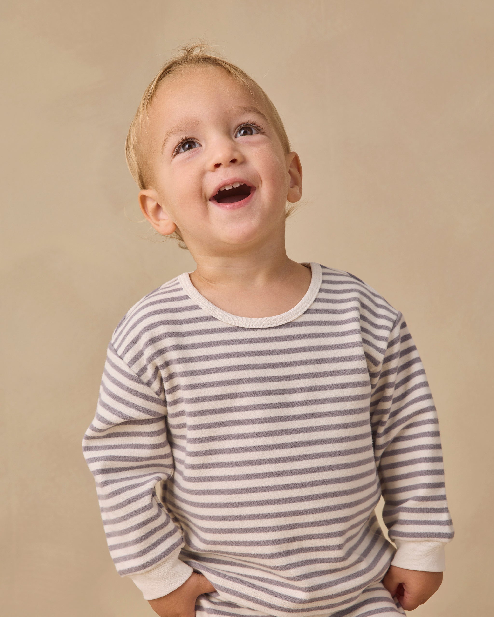 A young toddler with light hair looking up and smiling, wearing a long-sleeve shirt with thin gray horizontal stripes on an off-white background, set against a plain beige studio backdrop.