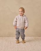A smiling toddler stands against a warm beige studio backdrop wearing a light beige and white checkered hooded jacket, gray pants, and tan shoes.