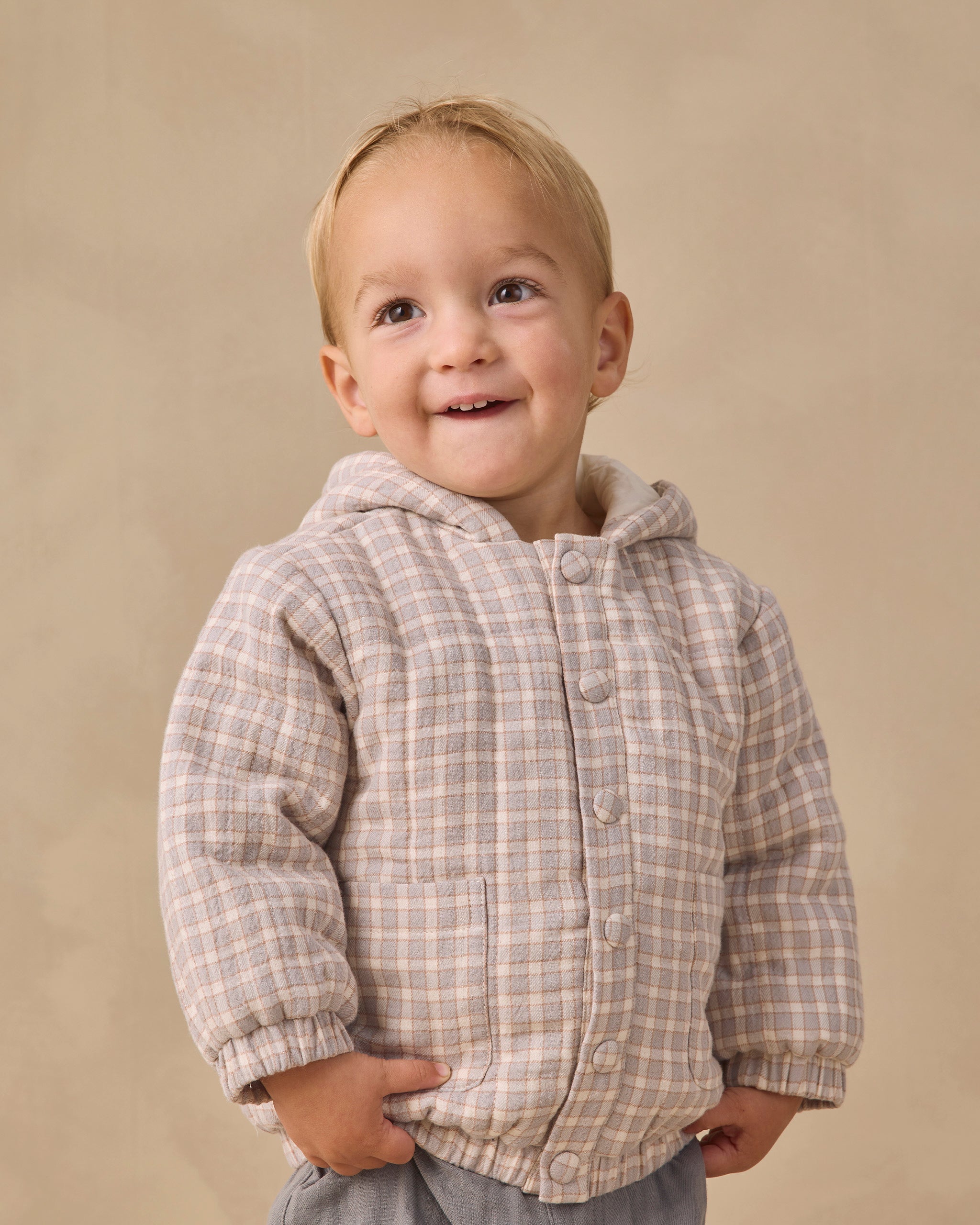 A smiling toddler wearing a beige and brown plaid hooded button-up jacket, standing against a solid beige studio background.