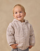 A smiling toddler wearing a beige and brown plaid hooded button-up jacket, standing against a solid beige studio background.