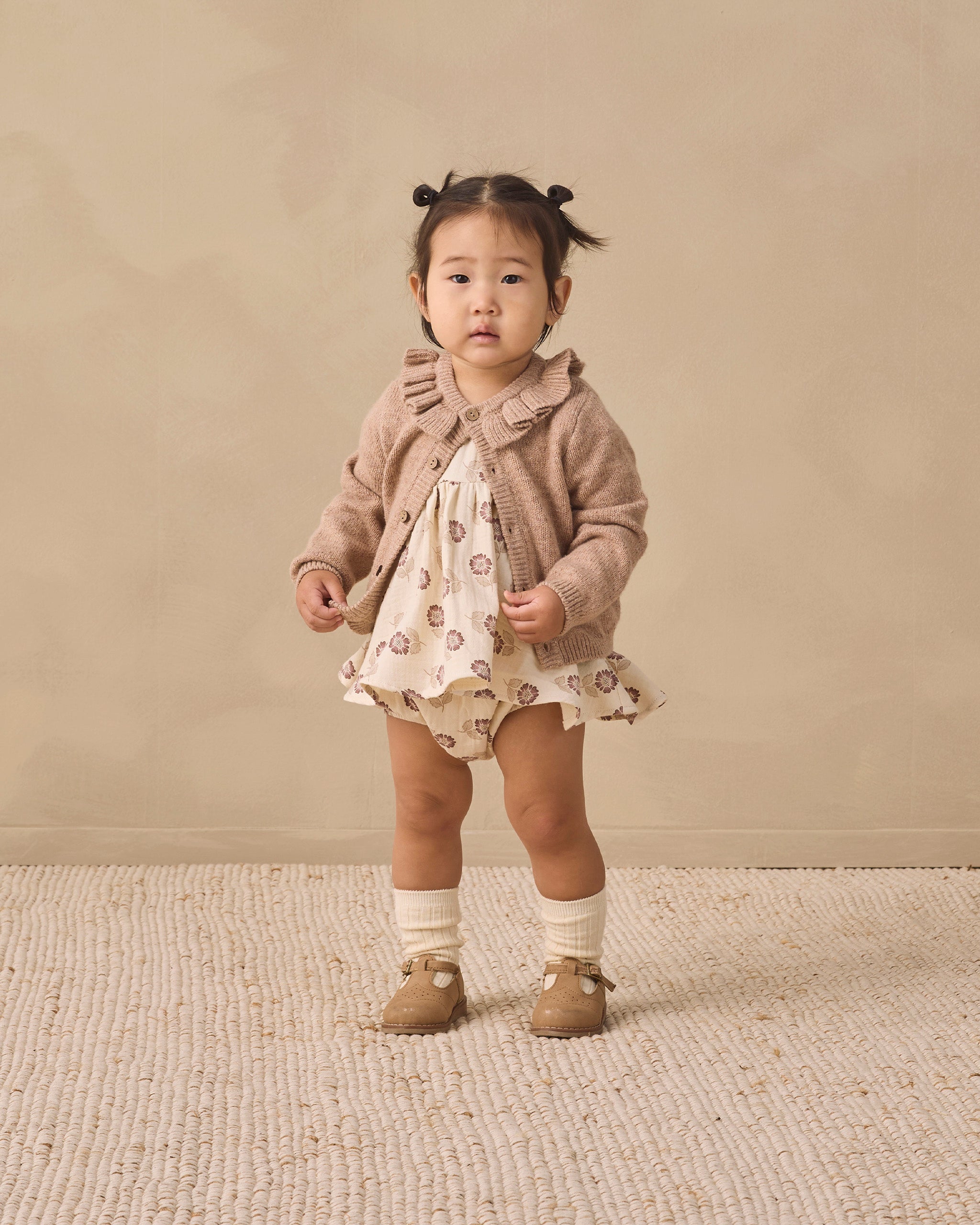A toddler stands against a warm beige studio backdrop wearing a cream dress with a brown animal print, a taupe ruffled cardigan, white socks, and tan shoes.