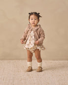 A toddler stands against a warm beige studio backdrop wearing a cream dress with a brown animal print, a taupe ruffled cardigan, white socks, and tan shoes.