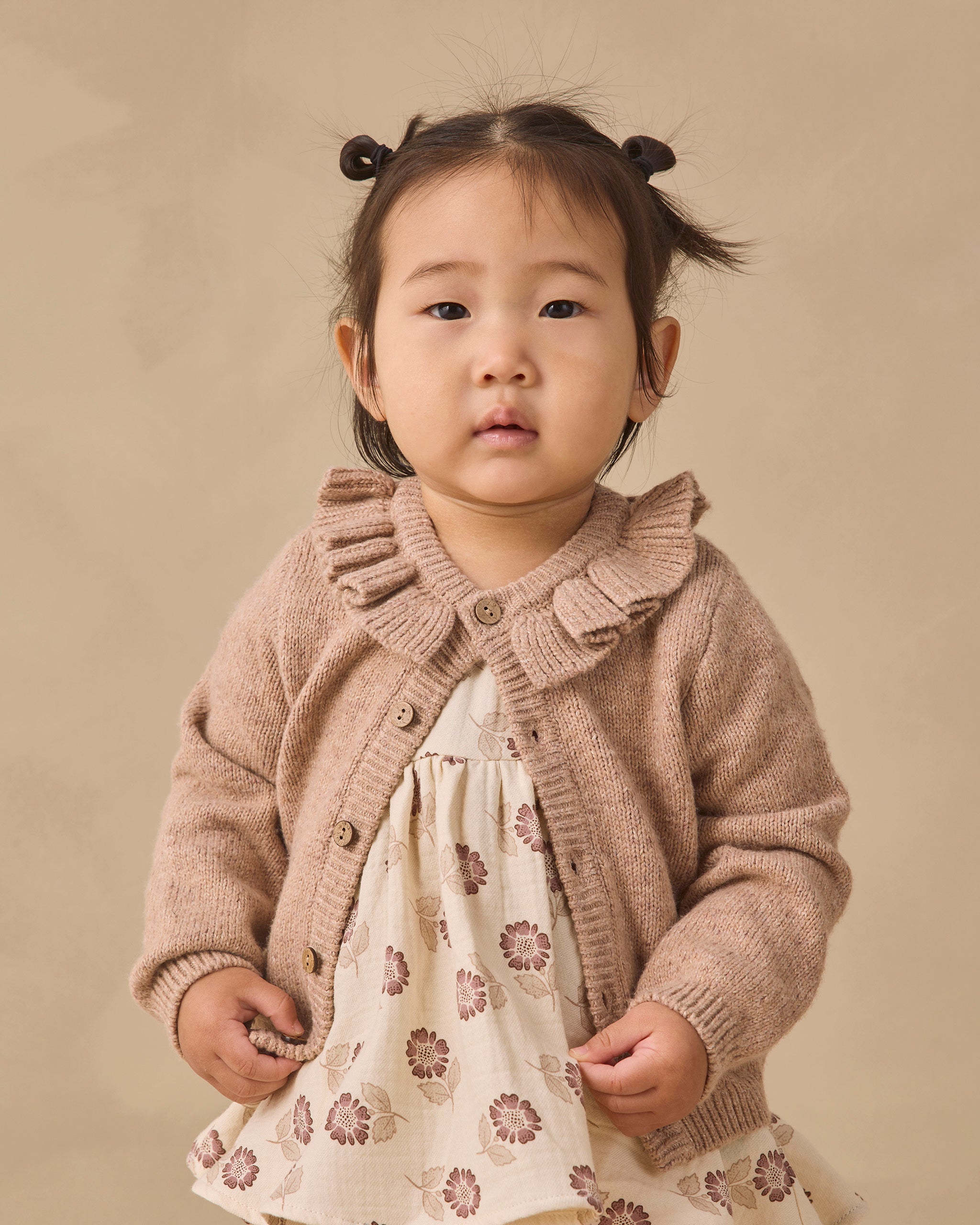 A toddler girl with dark hair in two buns wearing a beige knit cardigan with a ruffled collar over a cream dress with a brown floral pattern, standing against a solid beige background.