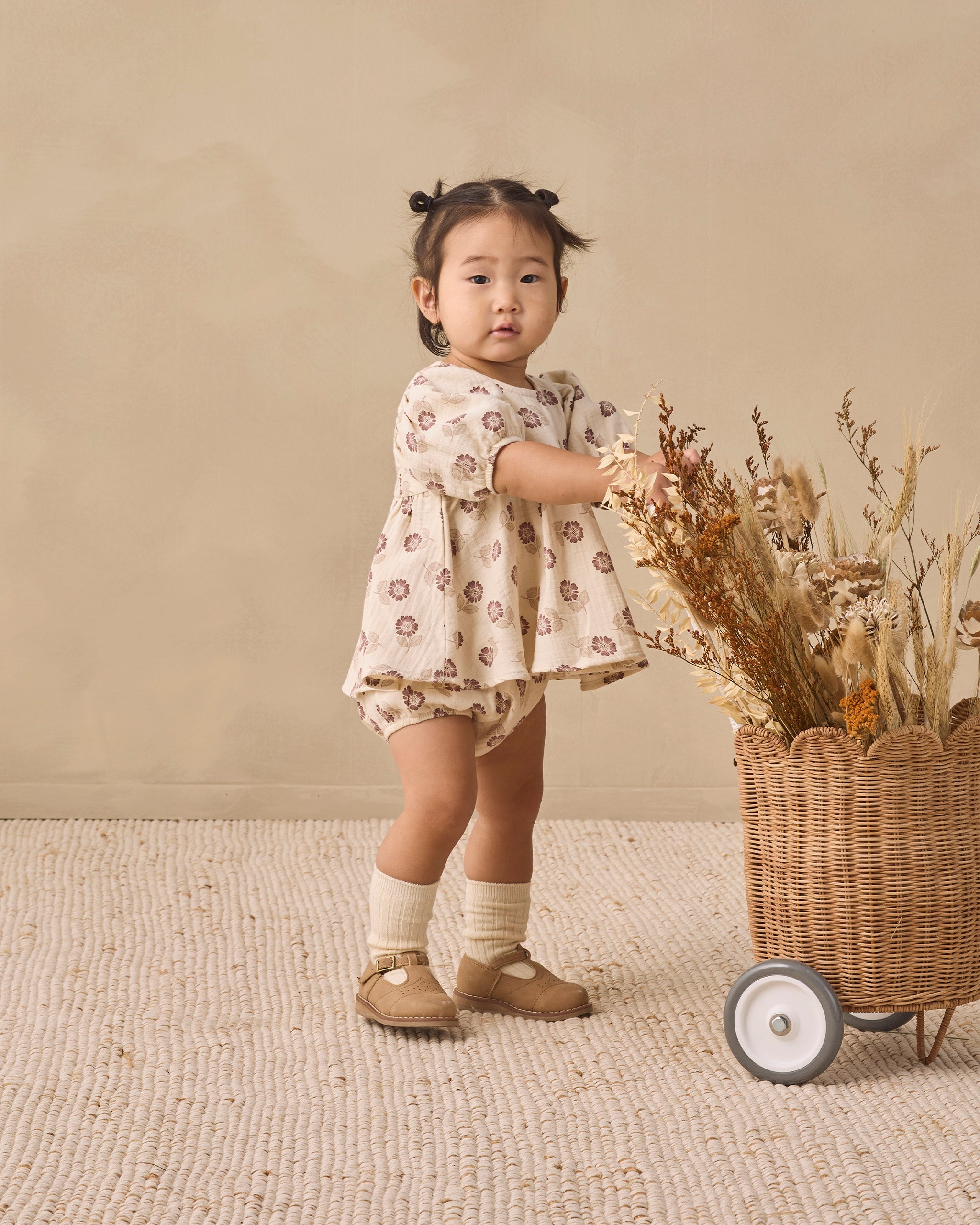A toddler girl stands in a studio setting wearing a cream-colored short-sleeve romper with a brown floral pattern, paired with cream socks and tan Mary Jane shoes. She is positioned next to a wicker basket filled with dried flowers against a neutral beige background.