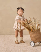 A toddler girl stands in a studio setting next to a wicker cart filled with dried flowers, wearing a cream short-sleeve romper with a brown floral pattern, cream socks, and tan shoes.