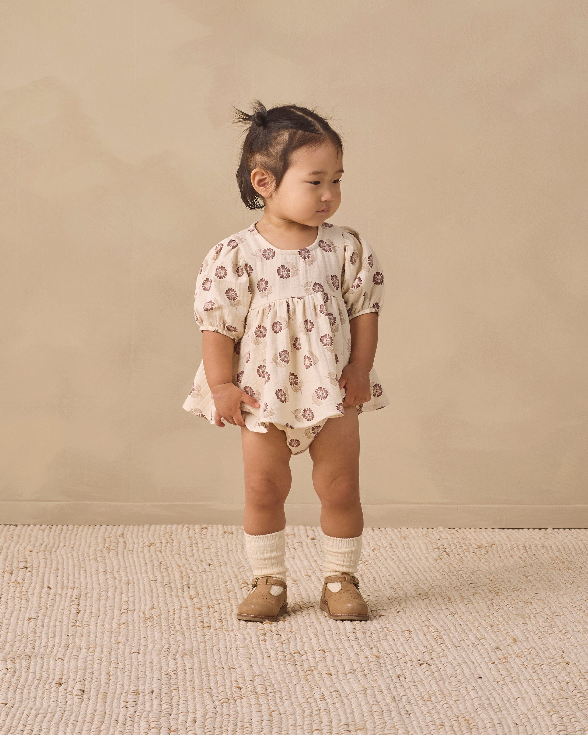 A toddler girl stands on a textured cream rug against a warm beige wall, wearing a cream short-sleeve romper with a brown floral pattern, cream knit socks, and tan shoes.