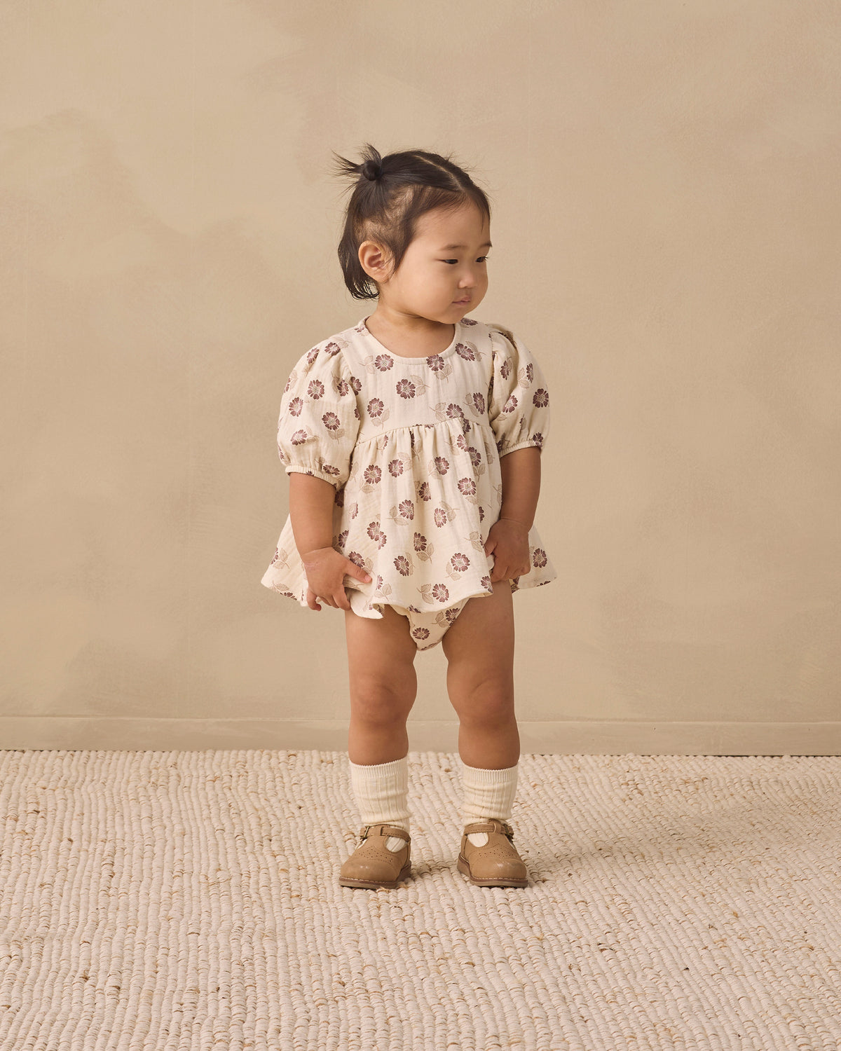 A toddler girl stands on a textured cream rug against a warm beige wall, wearing a cream short-sleeve romper with a brown floral pattern, cream knit socks, and tan shoes.