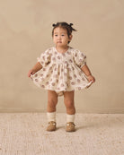 A toddler girl stands in a studio wearing a cream-colored puff-sleeve dress with a brown floral pattern, paired with cream socks and tan shoes, against a warm beige backdrop.