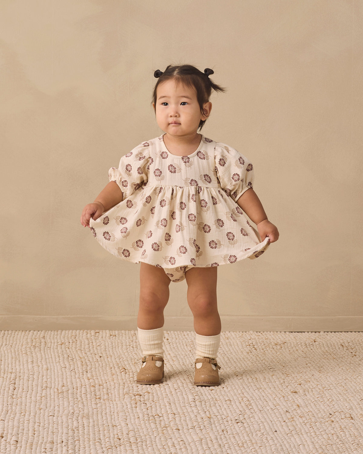 A toddler girl stands in a studio wearing a cream-colored puff-sleeve dress with a brown floral pattern, paired with cream socks and tan shoes, against a warm beige backdrop.