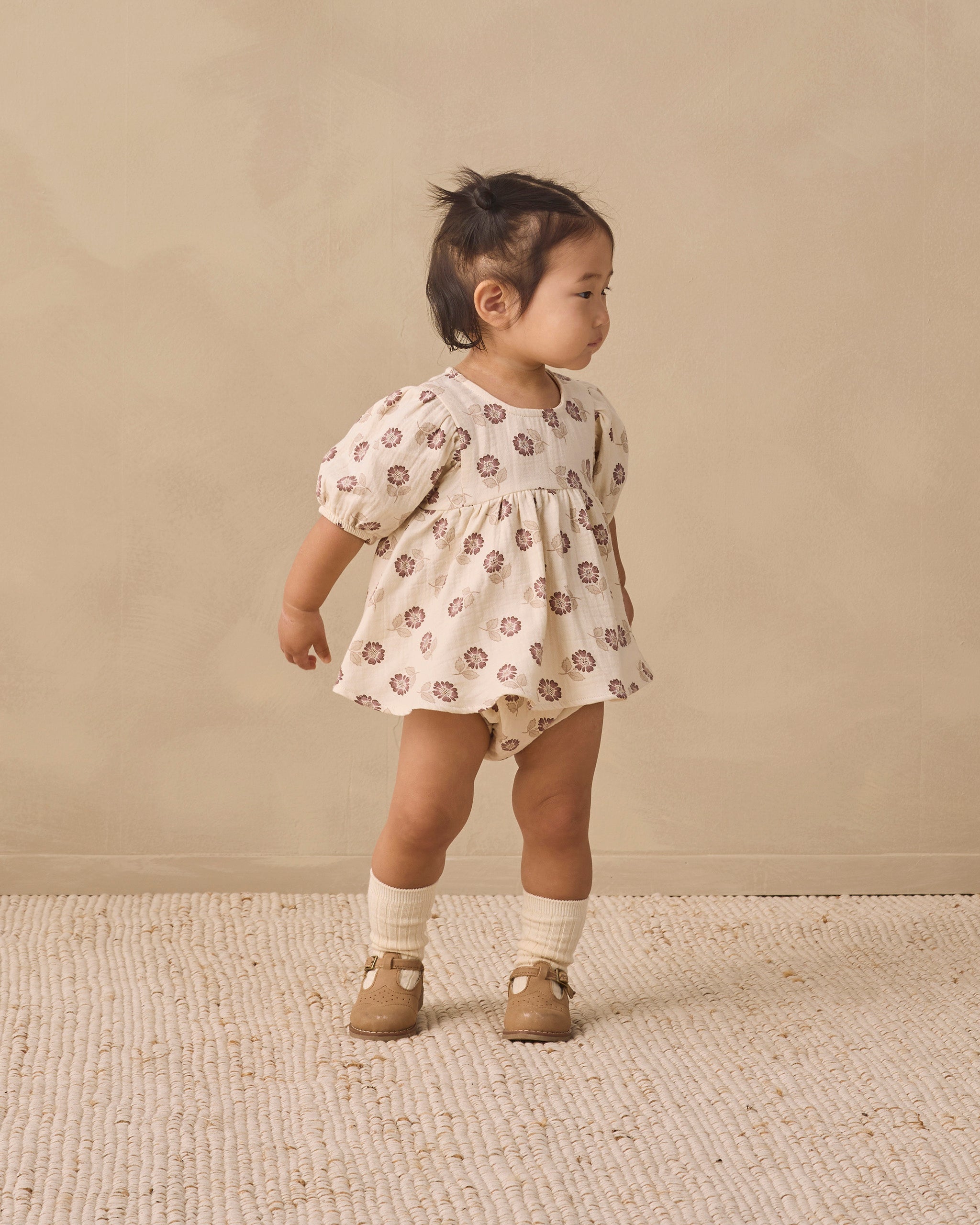 A toddler girl wearing a cream-colored short-sleeve romper with a brown floral pattern, standing on a textured beige rug against a neutral beige background.