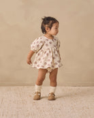 A toddler girl wearing a cream-colored short-sleeve romper with a brown floral pattern, standing on a textured beige rug against a neutral beige background.