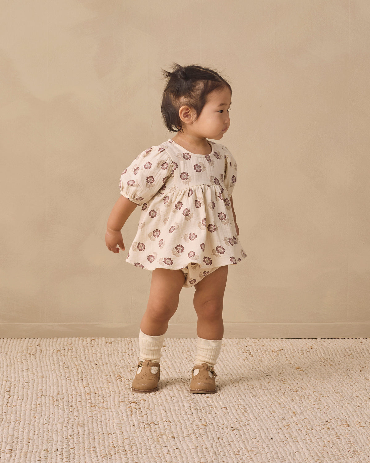 A toddler girl wearing a cream-colored short-sleeve romper with a brown floral pattern, standing on a textured beige rug against a neutral beige background.