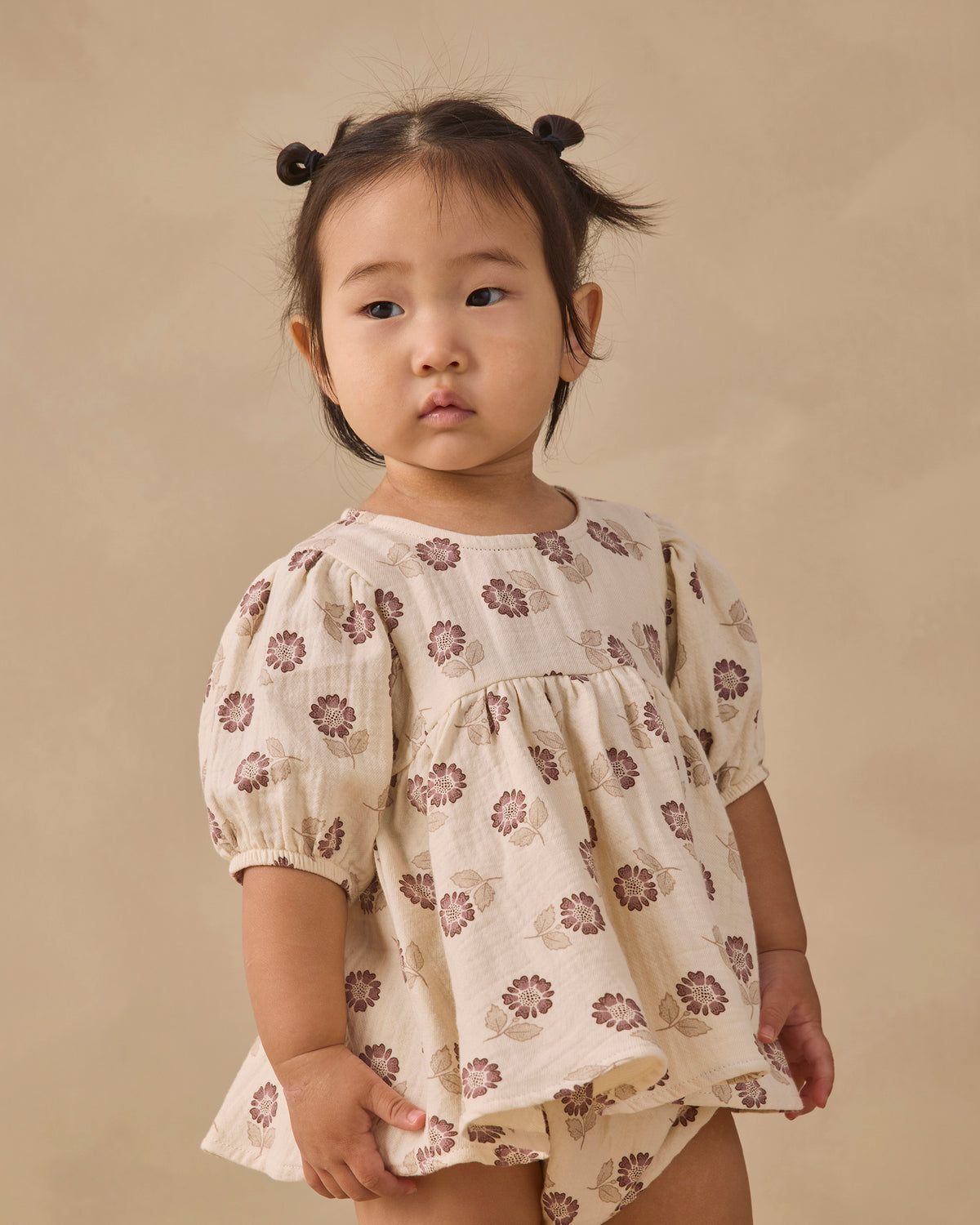A toddler girl with her hair in two buns wears a cream-colored short-sleeve dress with a brown floral pattern, standing against a solid beige background.