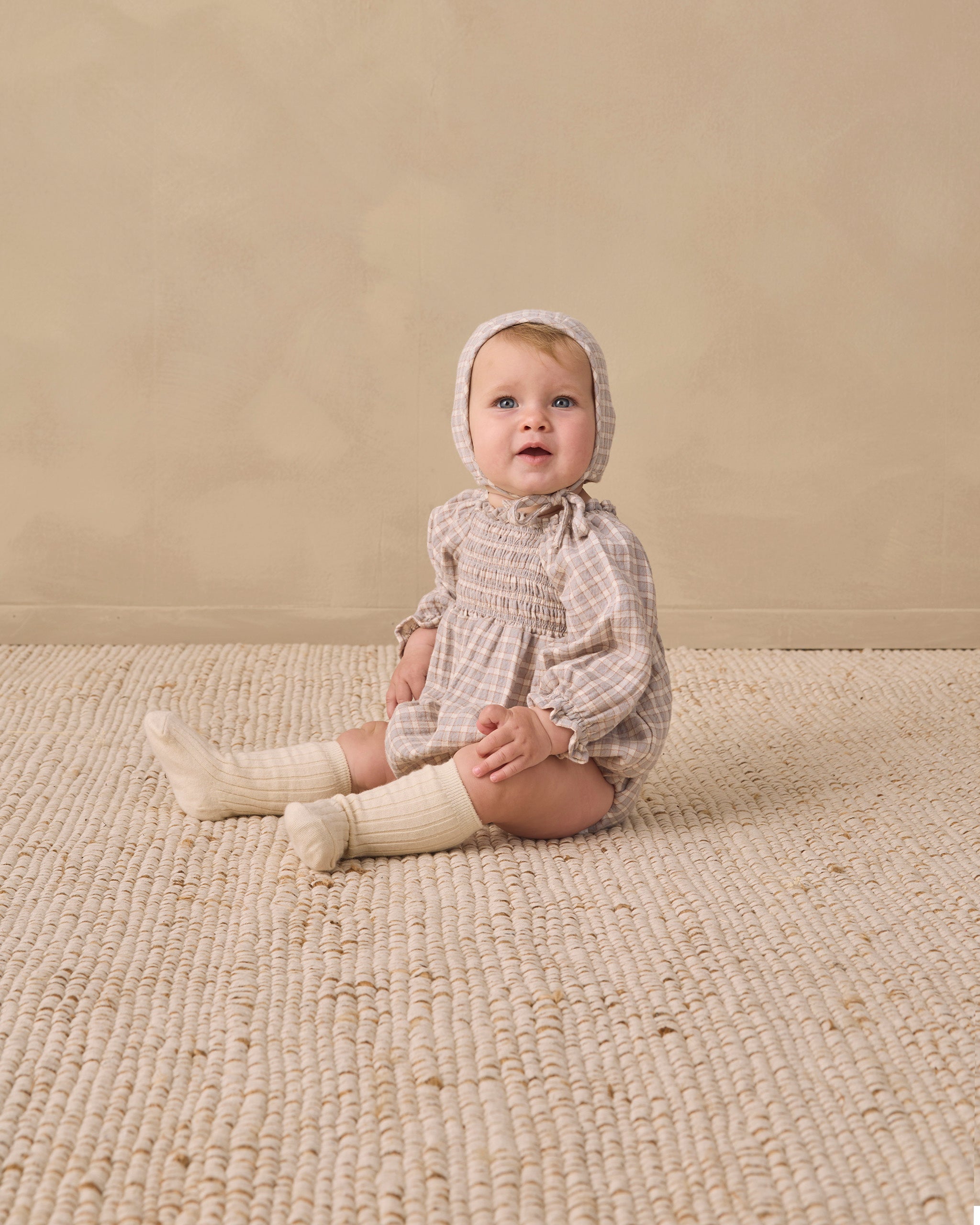 A baby sitting on a textured cream woven rug against a warm beige wall, wearing a beige and white checkered smocked romper, matching bonnet, and cream socks.