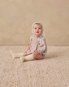 A baby sitting on a textured cream woven rug against a warm beige wall, wearing a beige and white checkered smocked romper, matching bonnet, and cream socks.