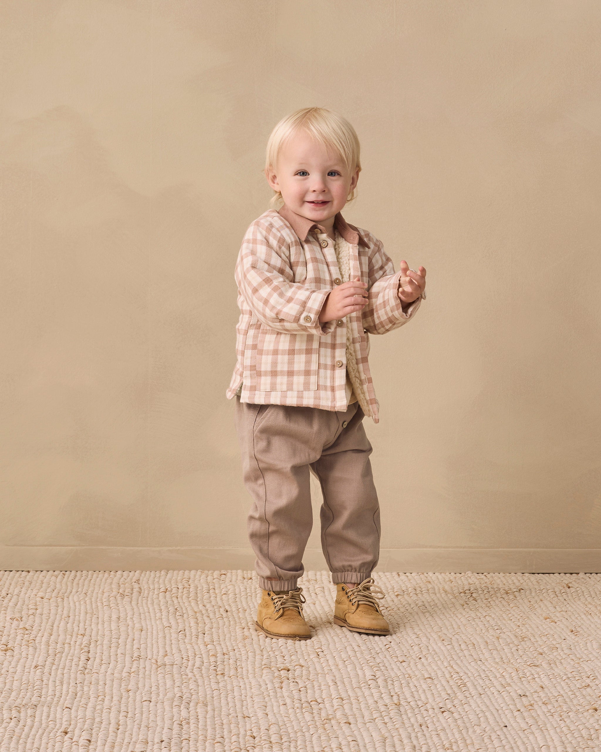 A toddler standing in a studio setting, wearing a beige and brown plaid button-up shirt, taupe pants, and tan boots against a warm beige backdrop.