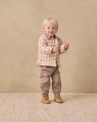 A toddler standing in a studio setting, wearing a beige and brown plaid button-up shirt, taupe pants, and tan boots against a warm beige backdrop.