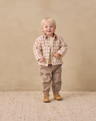 A toddler standing in a studio wearing a beige and brown plaid button-up shirt over a cream undershirt, paired with taupe pants and tan boots, against a warm beige backdrop.