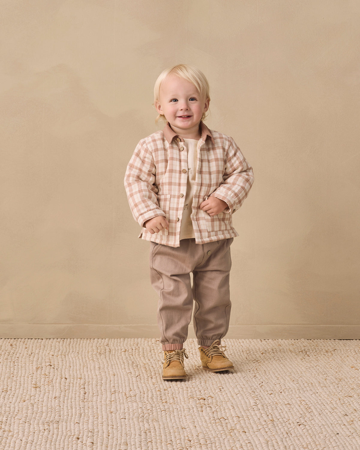 A toddler standing in a studio wearing a beige and brown plaid button-up shirt over a cream undershirt, paired with taupe pants and tan boots, against a warm beige backdrop.