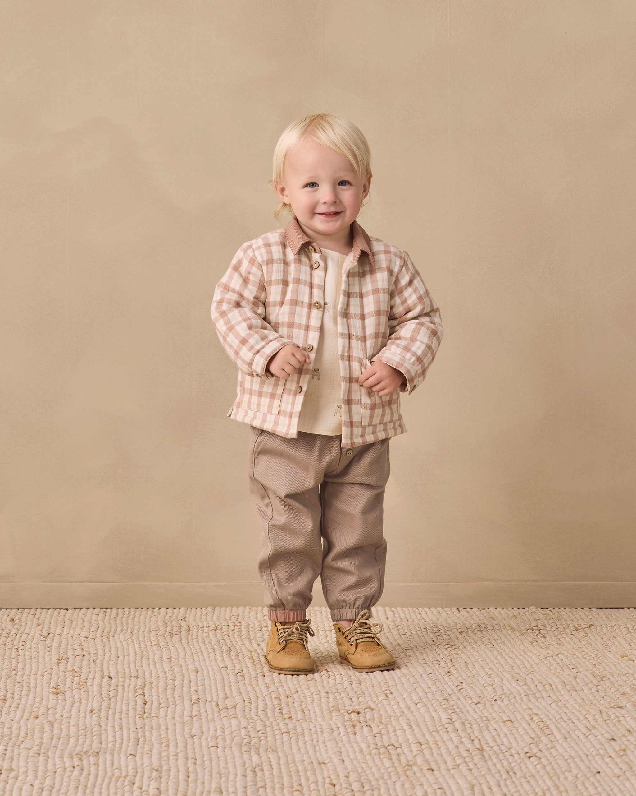 A toddler standing in a studio setting, wearing a beige and brown plaid button-up shirt over a cream undershirt, tan pants, and tan lace-up boots, against a warm beige backdrop.