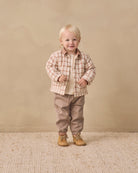 A toddler standing in a studio setting, wearing a beige and brown plaid button-up shirt over a cream undershirt, tan pants, and tan lace-up boots, against a warm beige backdrop.