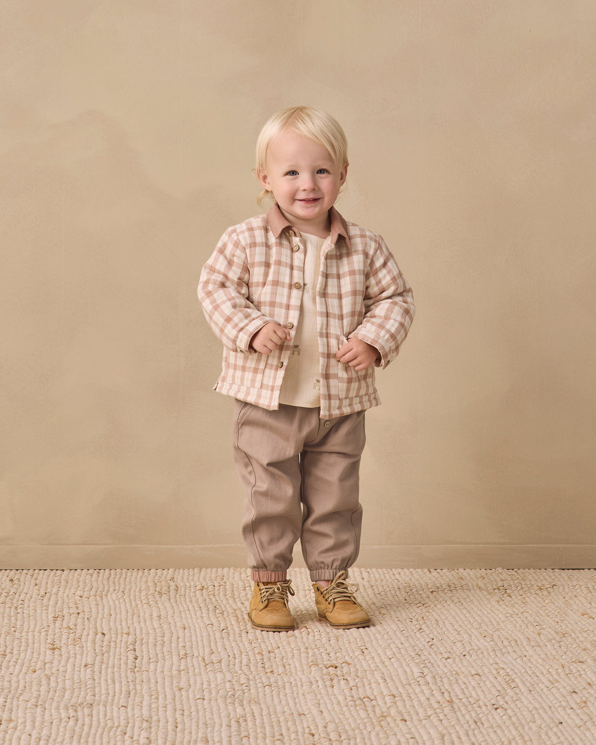 A toddler standing in a studio setting, wearing a beige and brown plaid button-up shirt over a cream undershirt, tan pants, and tan lace-up boots, against a warm beige backdrop.