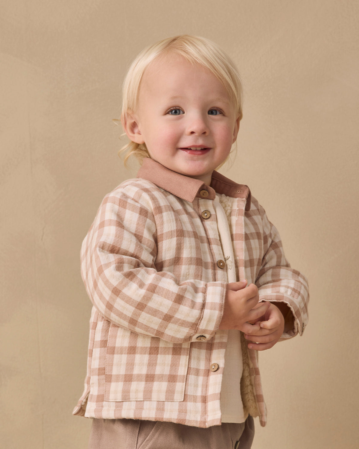 A smiling toddler with blonde hair wearing a beige and brown plaid button-up shirt jacket over a cream shirt, standing against a warm beige studio backdrop.