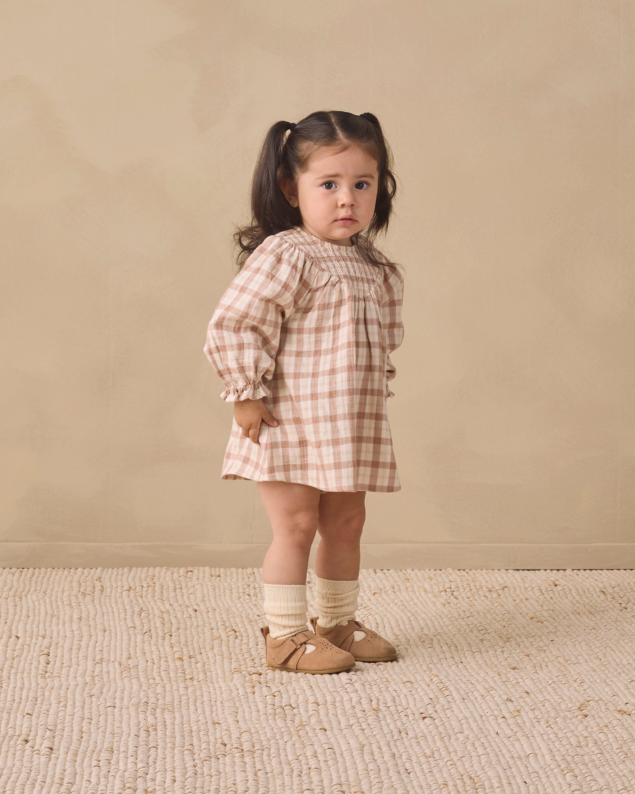 A young girl with dark hair in a ponytail stands in a studio wearing a beige and white gingham dress, cream socks, and tan shoes against a warm beige background.