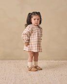 A young girl with dark hair in a ponytail stands in a studio wearing a beige and white gingham dress, cream socks, and tan shoes against a warm beige background.