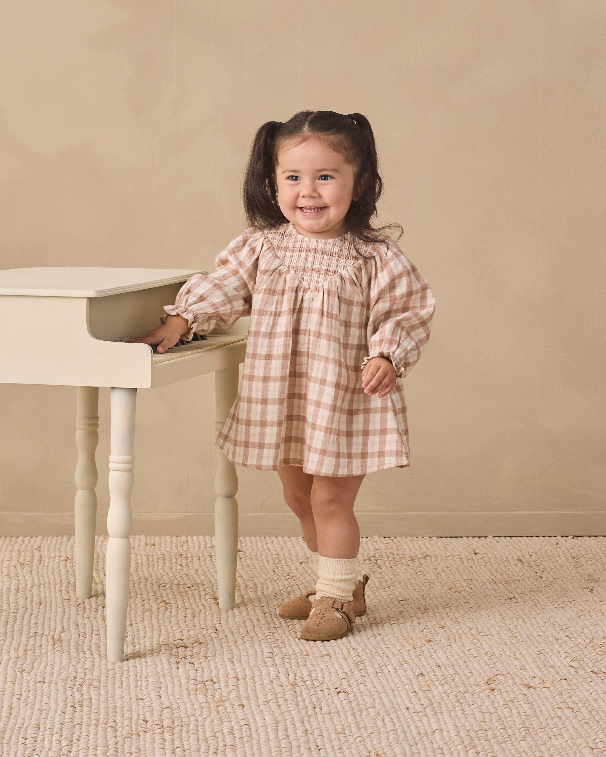 A young girl with dark hair in pigtails stands next to a small white table, wearing a beige and white gingham dress, cream socks, and tan shoes, against a neutral beige background.