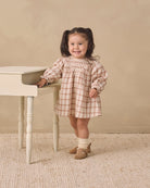 A young girl with dark hair in pigtails stands next to a small white table, wearing a beige and white gingham dress, cream socks, and tan shoes, against a neutral beige background.