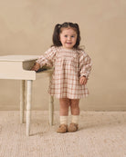 A young girl with dark hair in pigtails stands next to a small white piano, wearing a beige and white gingham dress, cream socks, and tan shoes, set against a neutral beige wall and textured rug.