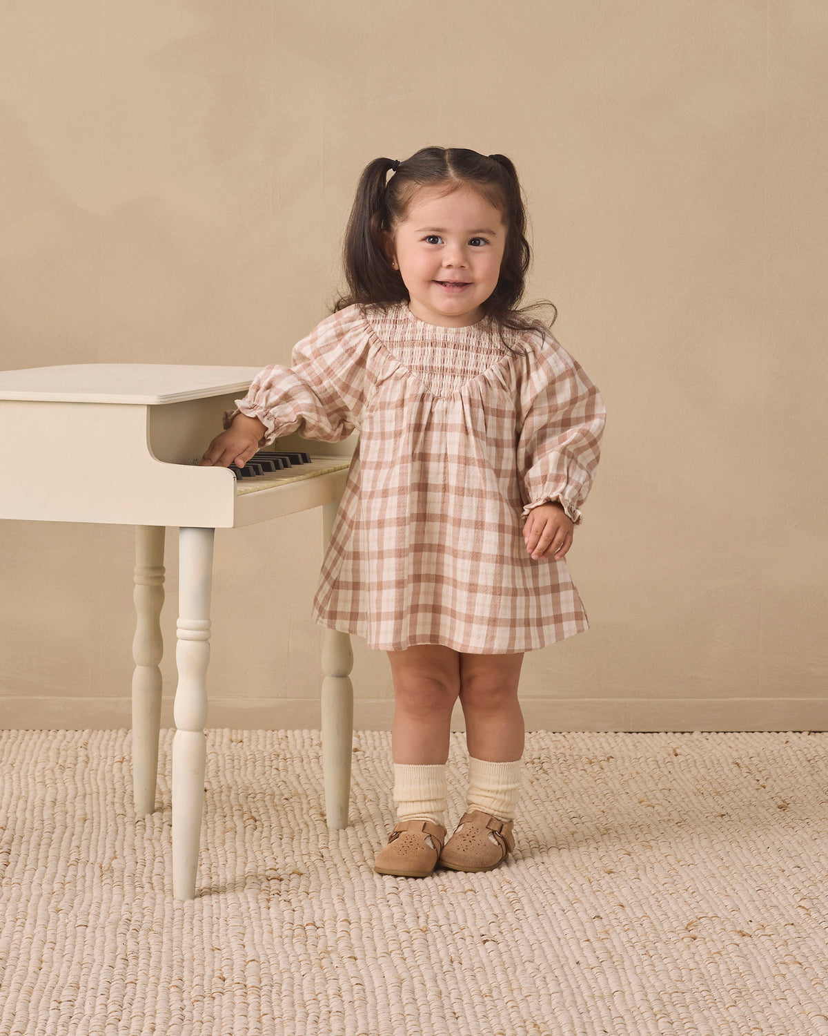 A young girl with dark hair in pigtails stands next to a small white piano, wearing a beige and white gingham dress, cream socks, and tan shoes, set against a neutral beige wall and textured rug.