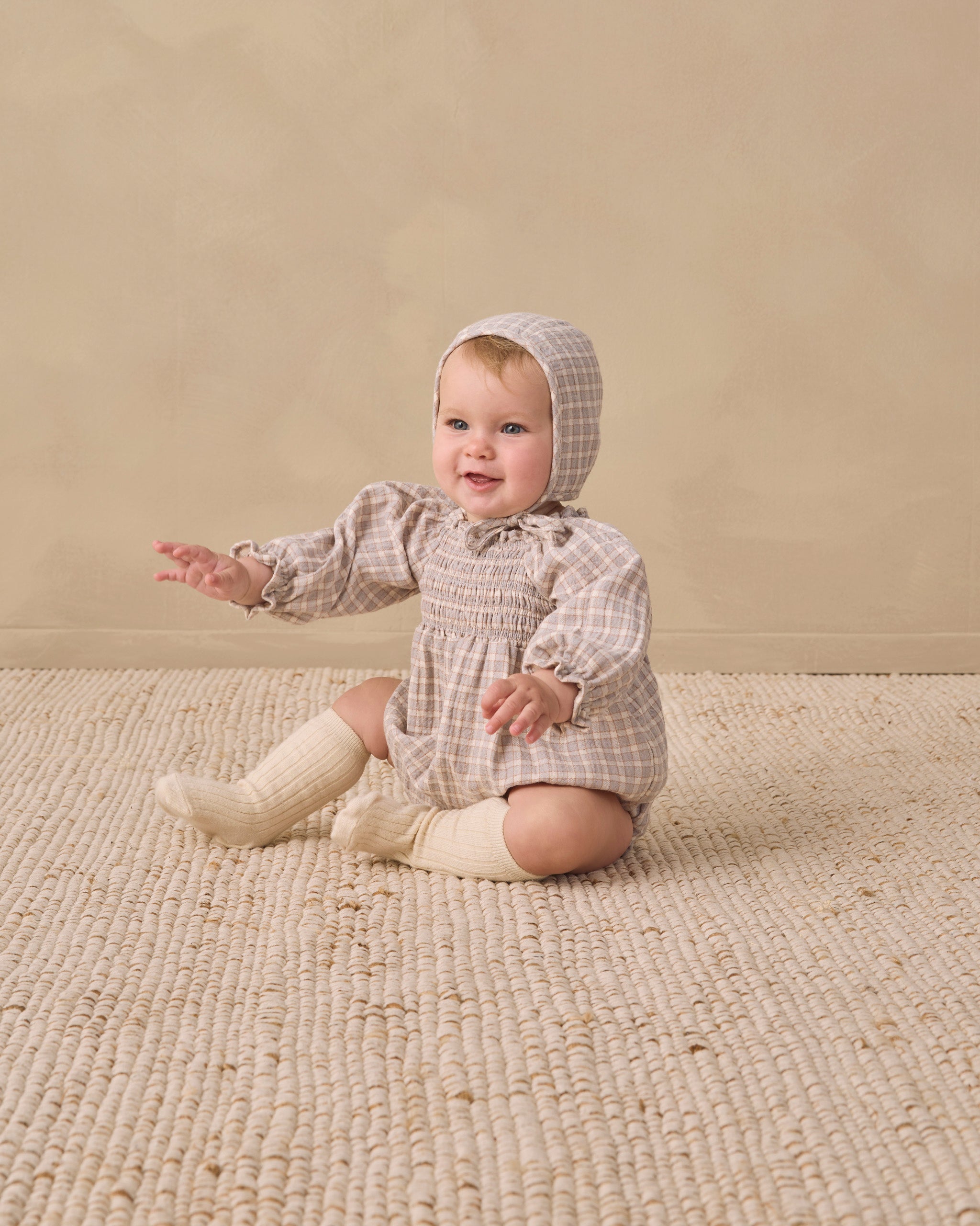 A baby sitting on a textured cream rug against a warm beige studio wall, wearing a beige and white checkered smocked romper with a matching bonnet and cream knit booties.