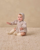 A baby sitting on a textured cream rug against a warm beige studio wall, wearing a beige and white checkered smocked romper with a matching bonnet and cream knit booties.
