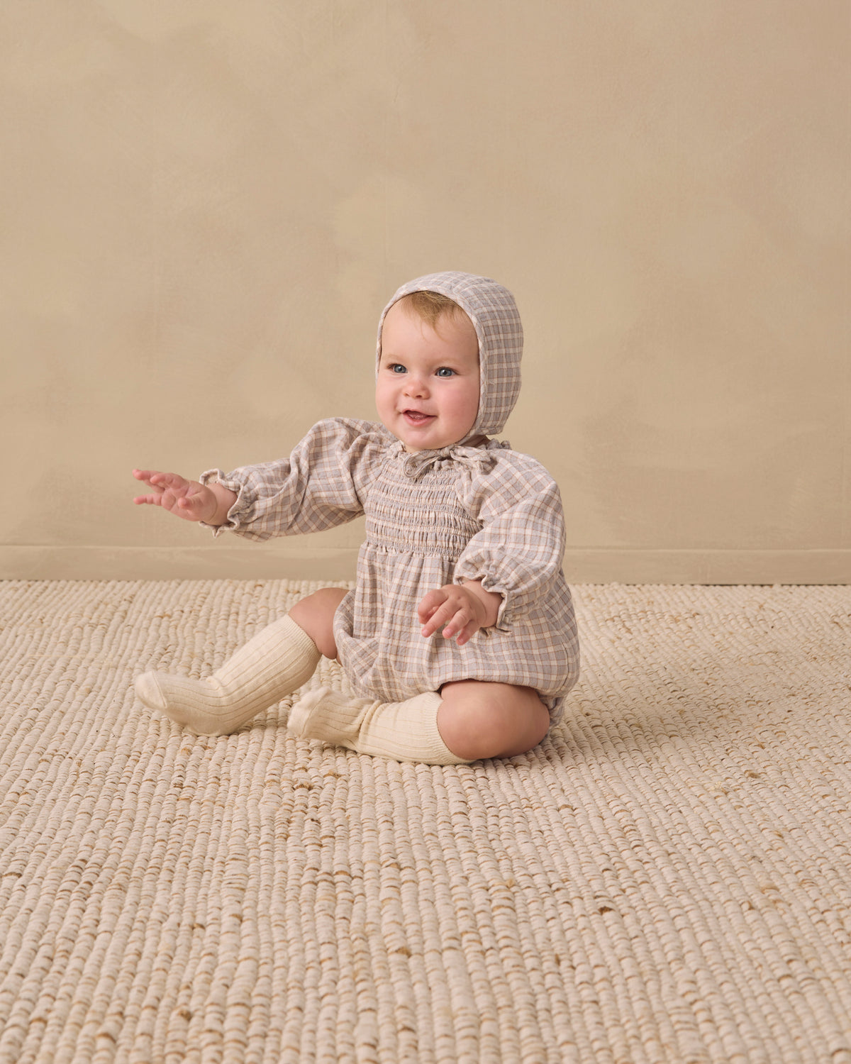 A baby sitting on a textured cream rug against a warm beige studio wall, wearing a beige and white checkered smocked romper with a matching bonnet and cream knit booties.