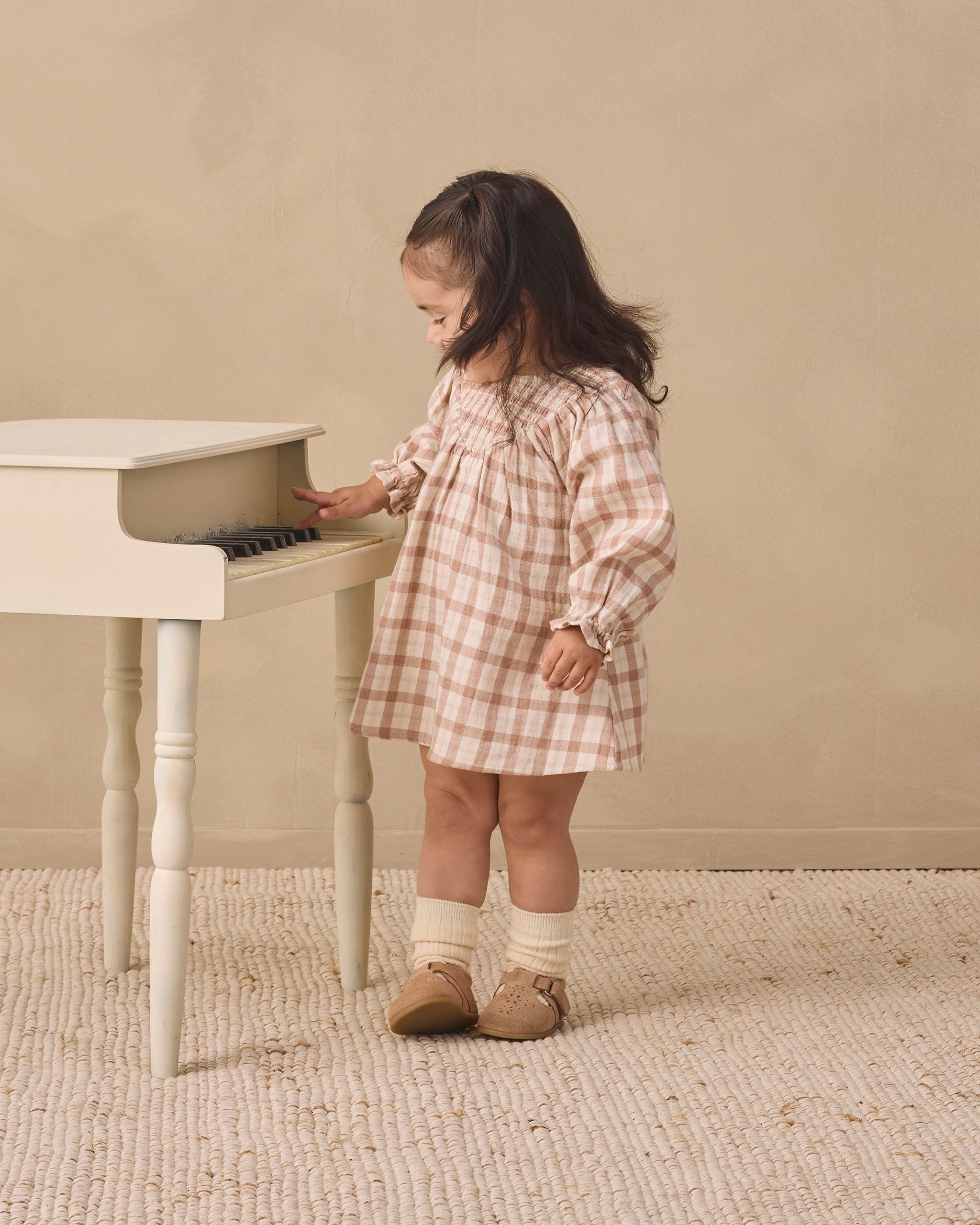 A toddler girl wearing a pink and beige gingham dress stands next to a small white piano, touching the keys. She is wearing cream socks and tan shoes in a warm, neutral-toned room with a textured rug.