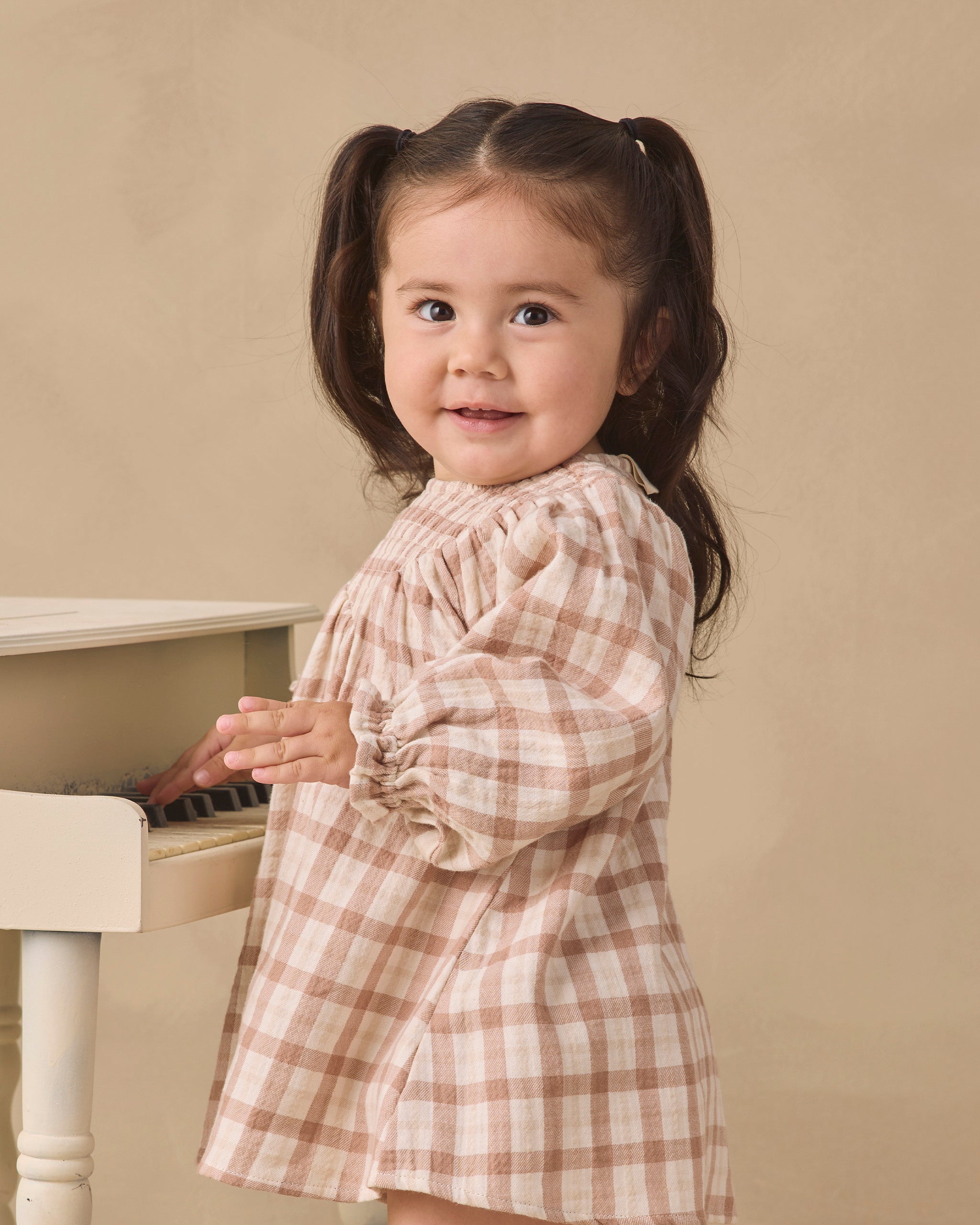 A young girl with dark brown pigtails stands next to a white piano, wearing a beige and brown plaid long-sleeve dress against a solid beige background.