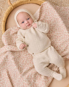 A baby wearing a cream-colored knit bonnet and matching footed sleeper lies on a pink and beige patterned blanket inside a woven rattan bassinet.
