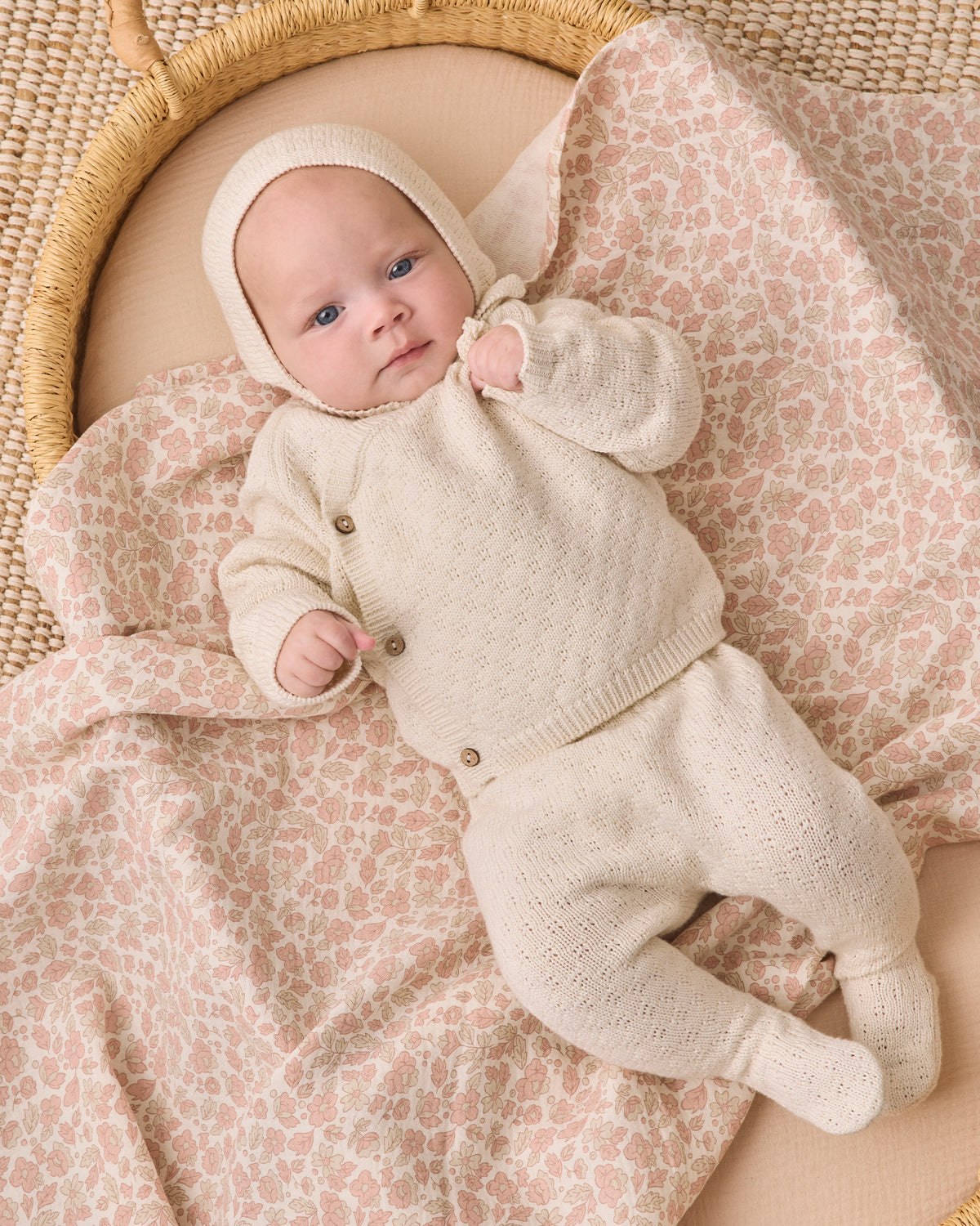 A baby wearing a cream-colored knit bonnet and matching footed sleeper lies on a pink and beige patterned blanket inside a woven rattan bassinet.