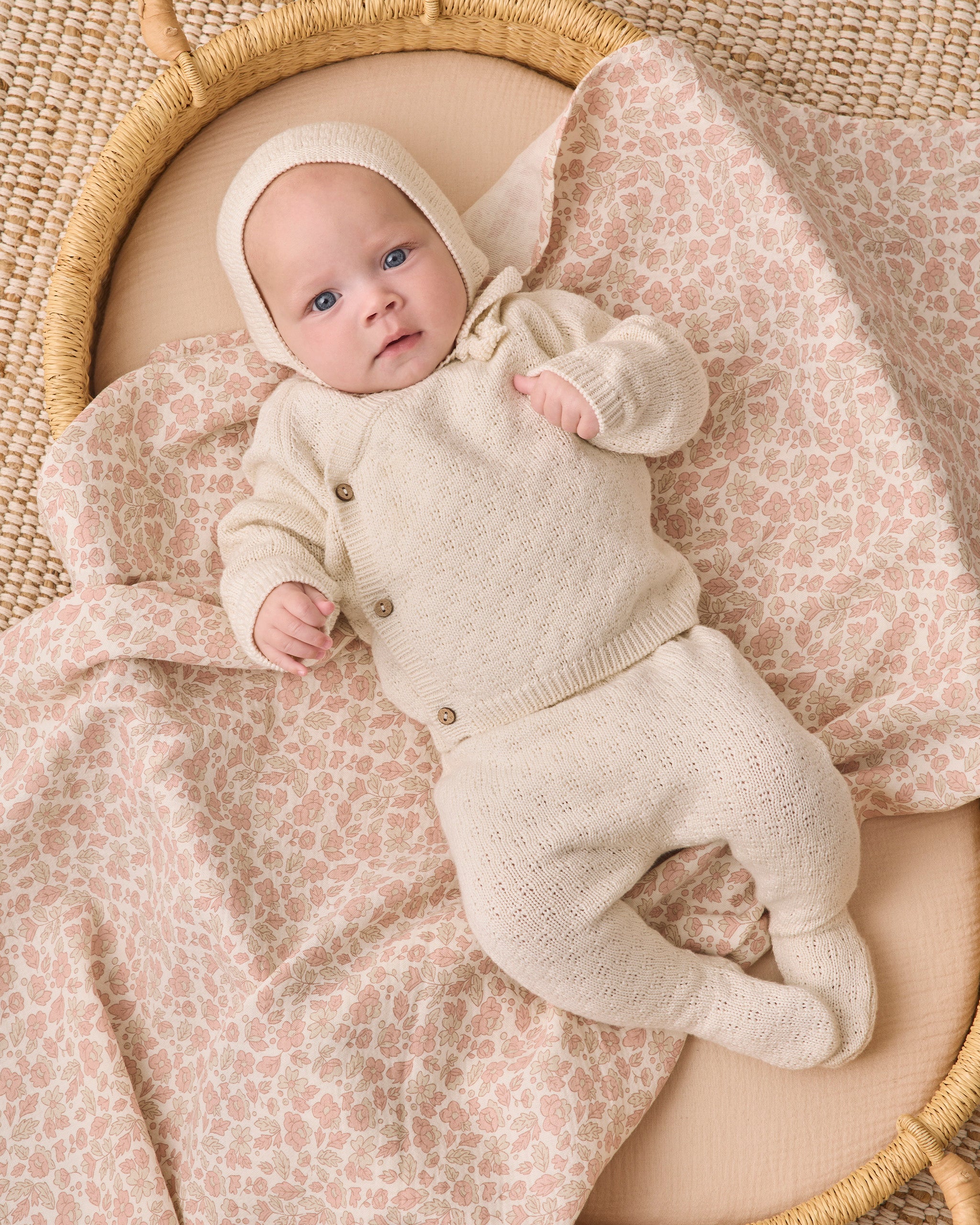 A baby lying on their back inside a woven basket, wearing a cream-colored knit one-piece outfit with a matching bonnet and wrapped in a beige and pink leopard print blanket.