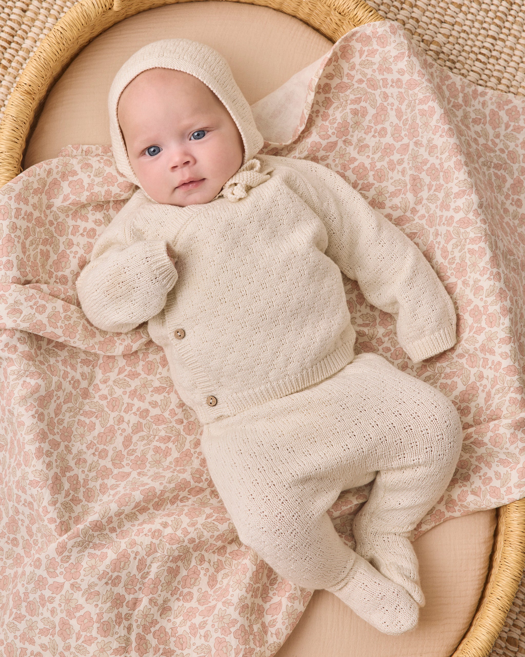 A baby wearing a cream-colored knit bonnet, cardigan, and footie sleeper set, lying on a pink and beige patterned blanket inside a woven bassinet.