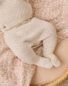 A close-up of a baby's footed knit romper in a warm cream or oatmeal color, styled on a pink and beige floral blanket.