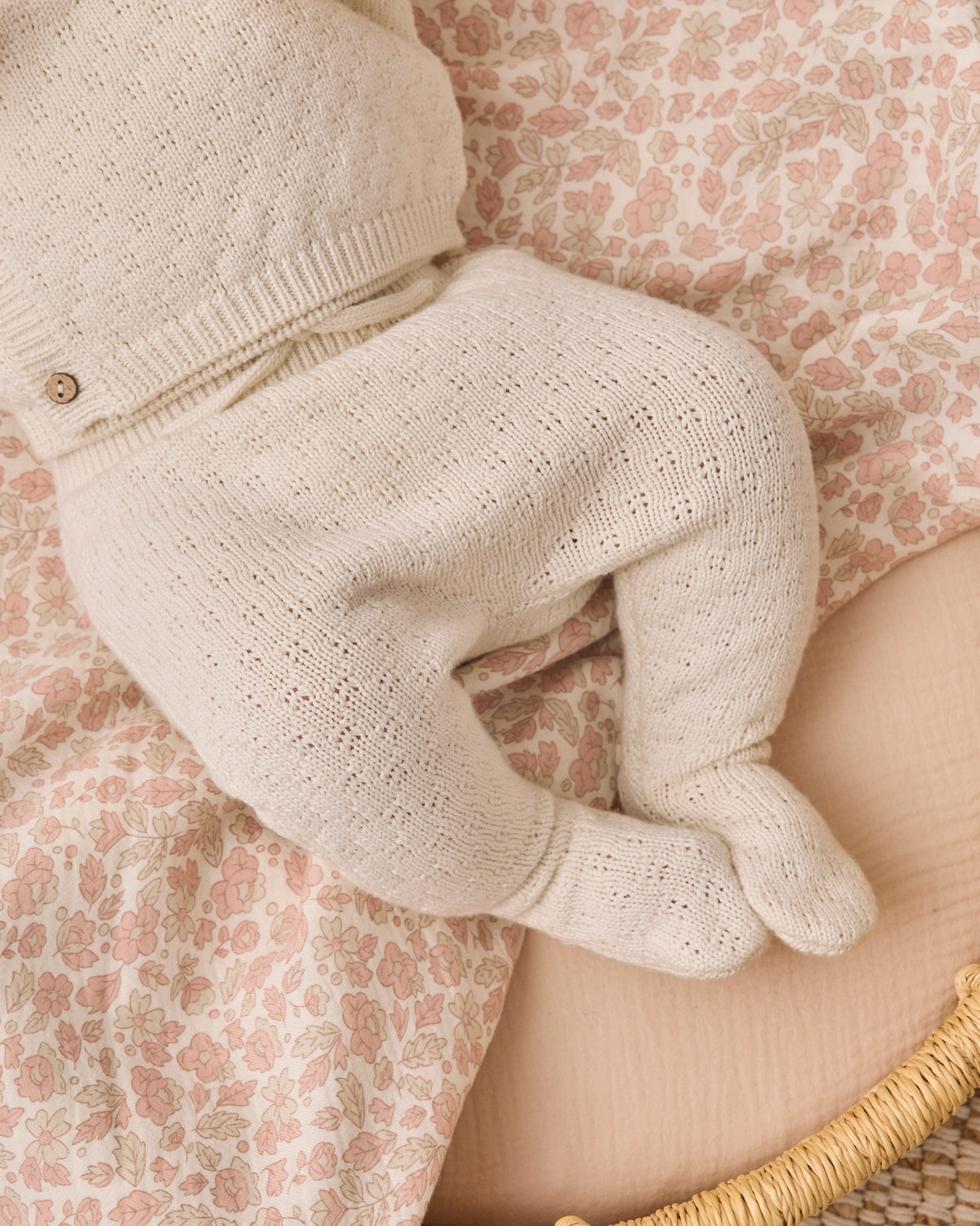 A close-up of a baby's footed knit romper in a warm cream or oatmeal color, styled on a pink and beige floral blanket.