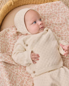 A baby wearing a cream-colored knit bonnet and matching cardigan lies on a patterned blanket inside a woven basket.